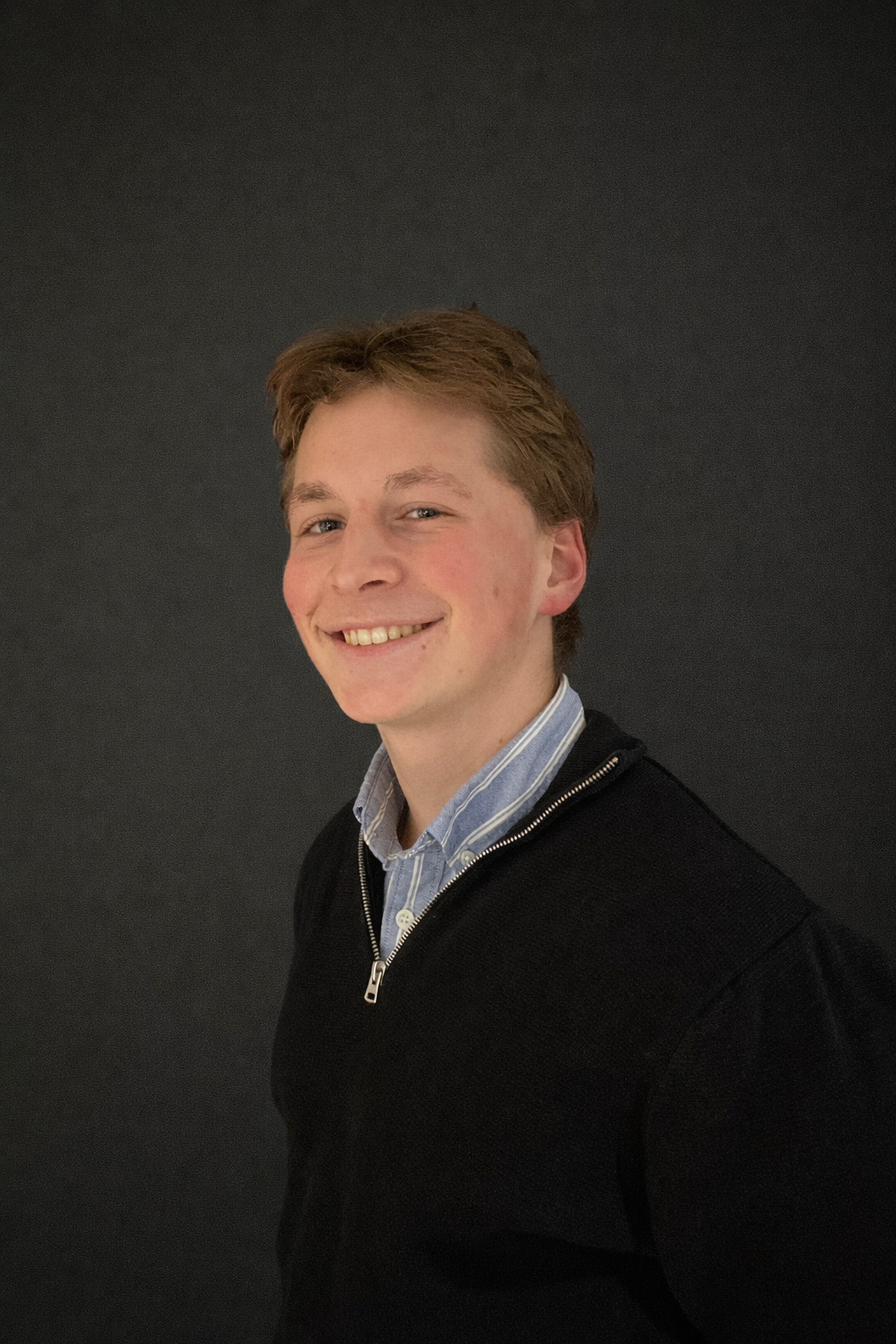 Een jongeman met kort lichtbruin haar glimlacht naar de camera, draagt een zwarte trui met rits over een blauw hemd met kraag, voor een effen donkere achtergrond-Daan van der Velden.